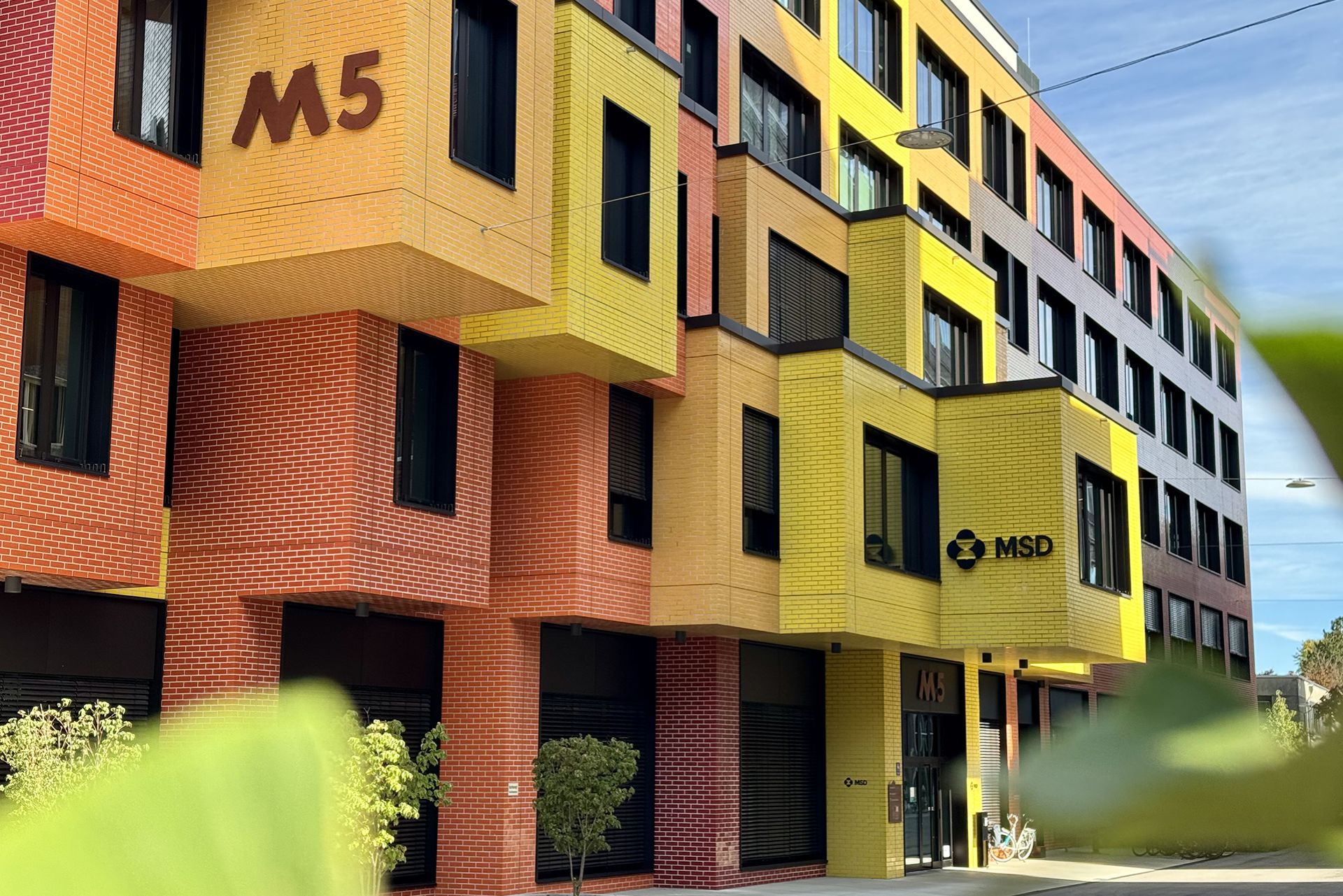 Buntes, modernes Bürogebäude mit der Aufschrift „MSD“ und „M5“ an der Fassade. Die Fassade ist in leuchtenden Gelb-, Orange- und Rottönen gehalten. Vor dem Gebäude stehen kleine Bäume und Fahrräder, was eine offene und urbane Umgebung signalisiert.
