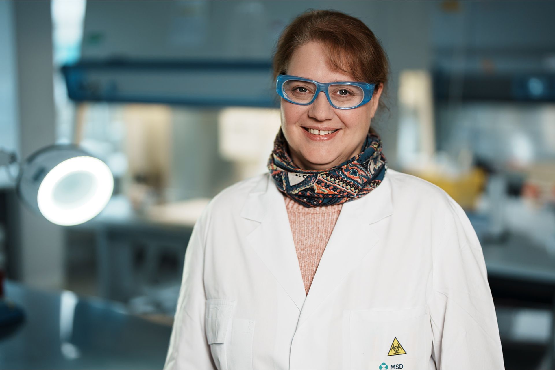 Eine Frau in einem Labor trägt einen weißen Laborkittel, eine bunte gemusterte Halsschleife und eine blaue Schutzbrille. Sie lächelt in die Kamera. Im Hintergrund ist ein Laborarbeitsplatz mit Beleuchtung und technischen Geräten zu sehen. Die Atmosphäre wirkt professionell und freundlich.