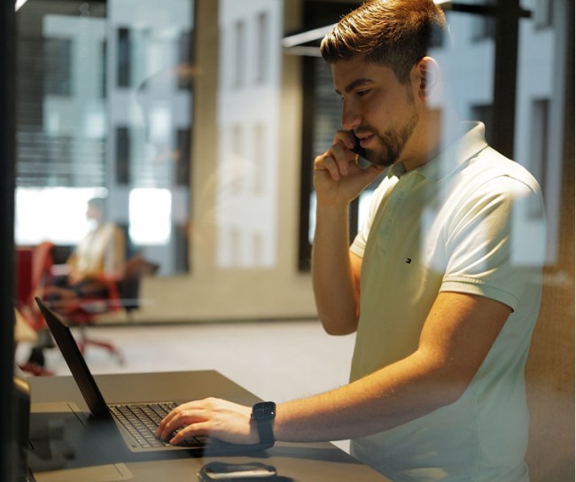 Ein junger Mann mit kurzen Haaren und Bart steht an einem Stehschreibtisch in einem modernen Büro. Er trägt ein helles Poloshirt, telefoniert mit einem Smartphone und arbeitet gleichzeitig an einem Laptop. Durch eine Glaswand sind unscharf weitere Personen im Hintergrund erkennbar.