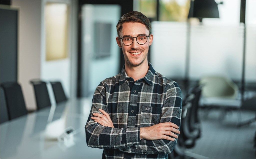 Ein junger Mann mit Brille und kariertem Hemd steht lächelnd mit verschränkten Armen in einem modernen Konferenzraum. Der Hintergrund zeigt einen Besprechungstisch und Glaswände, die eine professionelle, aber freundliche Atmosphäre vermitteln.