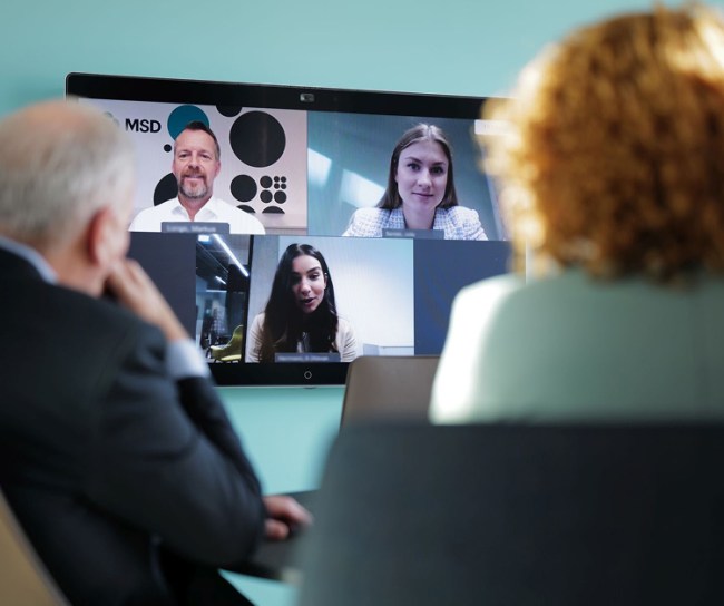 Zwei Personen sitzen vor einem großen Bildschirm und führen ein virtuelles Meeting mit vier anderen Teammitgliedern.