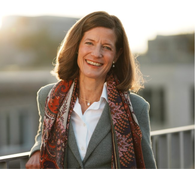 Porträt einer Frau mit braunen Haaren, Blazer und gemustertem Schal, die bei Sonnenuntergang auf einer Dachterrasse steht und lächelt.
