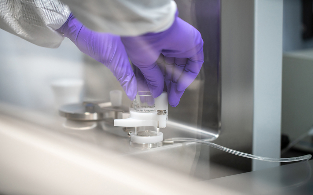 Eine Person mit violetten Schutzhandschuhen arbeitet an einem präzisen technischen Gerät in einer sterilen Laborumgebung. Die Hände positionieren ein kleines Glasgefäß in eine Vorrichtung, vermutlich zur Messung oder Analyse. Die Szene vermittelt hohe Sorgfalt und Laborpräzision.