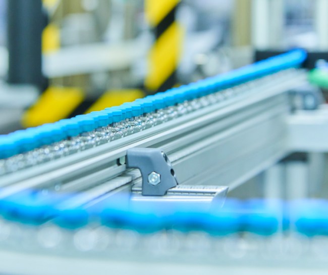 Nahaufnahme einer automatisierten Förderstrecke mit kleinen Glasbehältern, die in blauen Halterungen transportiert werden. Die Szene vermittelt Präzision und moderne Produktionstechnologie.