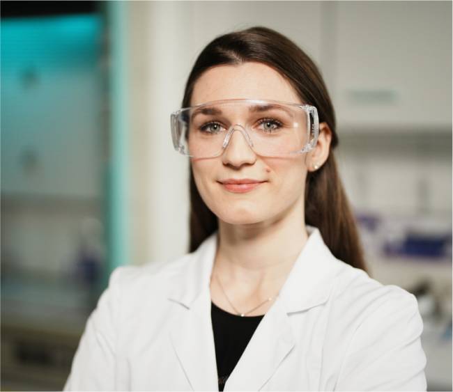 Eine Frau im weißen Laborkittel steht mit verschränkten Armen in einem Labor. Sie trägt eine Schutzbrille und lächelt leicht in die Kamera. Im Hintergrund ist Laborequipment zu sehen, was Professionalität und Selbstbewusstsein vermittelt.