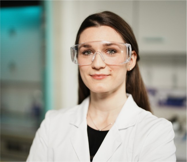 Eine Frau im weißen Laborkittel steht mit verschränkten Armen in einem Labor. Sie trägt eine Schutzbrille und lächelt leicht in die Kamera. Im Hintergrund ist Laborequipment zu sehen, was Professionalität und Selbstbewusstsein vermittelt.