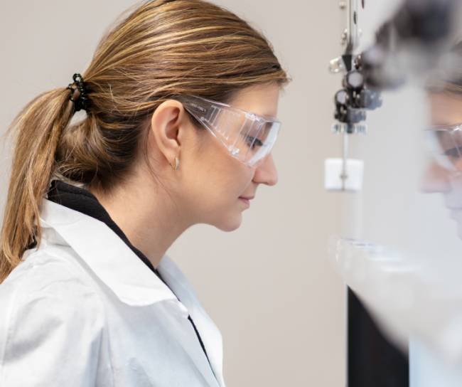 Eine Frau in Laborkittel und Schutzbrille beobachtet konzentriert ein Analysegerät oder eine Prüfanlage. Die Umgebung wirkt klinisch sauber, was auf einen Qualitätskontrollprozess hinweist.