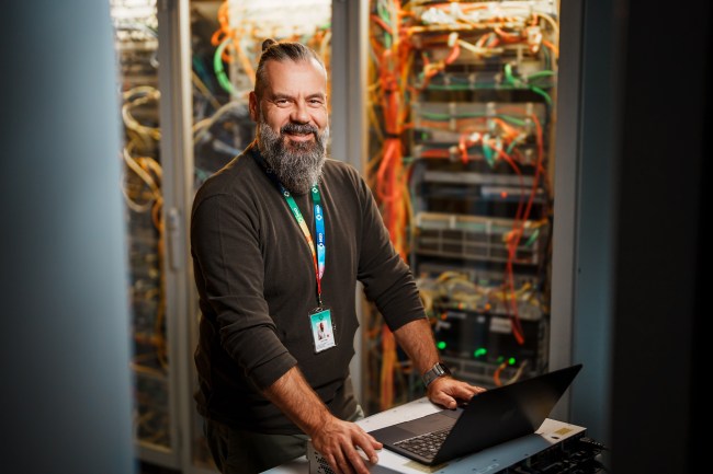Ein IT-Mitarbeiter mit Bart und langem Haar steht lächelnd vor einem geöffneten Serverrack voller bunter Kabel. Er trägt einen Mitarbeiterausweis und arbeitet an einem Laptop auf einem Rollwagen. Die Szene vermittelt technische Kompetenz und eine freundliche Arbeitsatmosphäre im Serverraum.