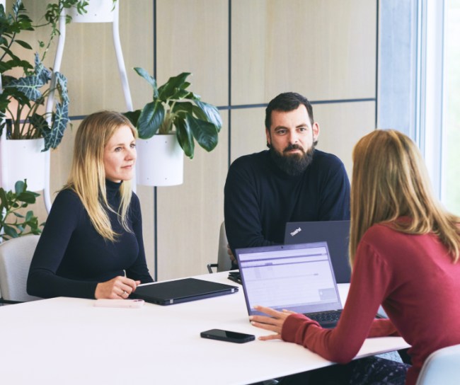 Drei Personen in einem Besprechungsraum diskutieren konzentriert an einem Tisch mit Laptops und Dokumenten.