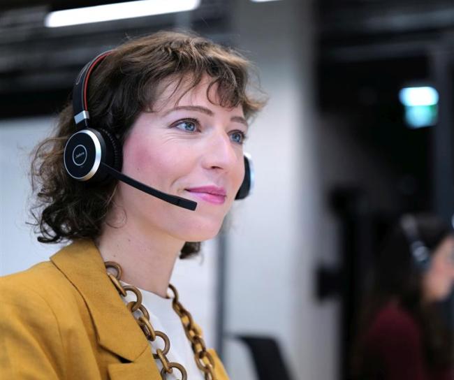 Eine junge Frau mit Headset und Brille sitzt an einem Arbeitsplatz im Großraumbüro. Sie lächelt und scheint sich in einem Gespräch zu befinden. Die Umgebung ist modern und hell, was auf ein dynamisches Arbeitsumfeld schließen lässt.