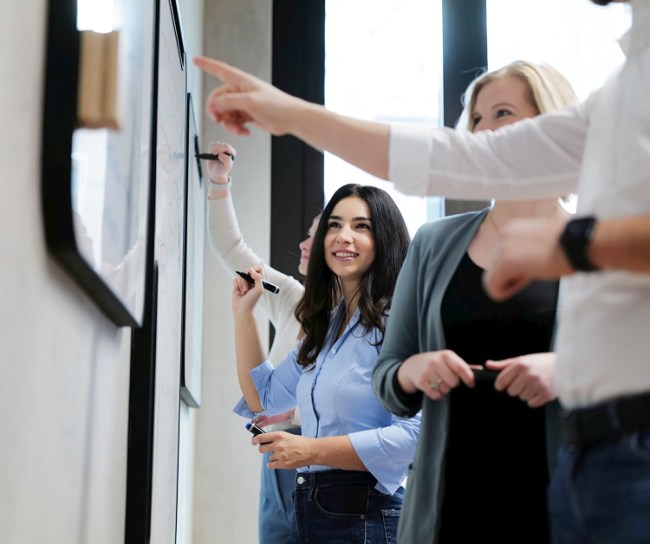 Mehrere junge Menschen arbeiten gemeinsam an einem Whiteboard. Eine Frau im Vordergrund lächelt, während andere diskutieren und zeigen. Die Szene zeigt Teamarbeit und kreative Zusammenarbeit in einem modernen Büro.